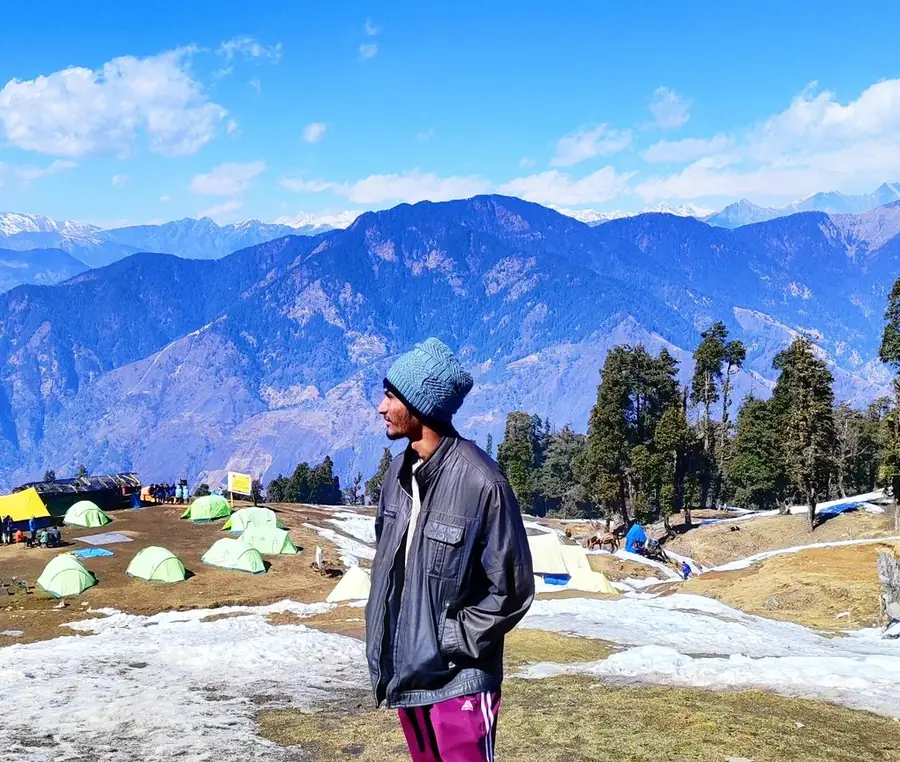 Manan Shah in the Himalayas, in front of a campsite and snow-capped mountains